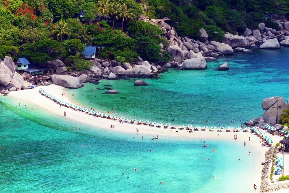 ภาพมุมสูงของชายหาดเขตร้อน เที่ยวสุราษฎร์ธานี ในสุราษฎร์ธานีที่เที่ยว มีน้ำทะเลสีฟ้าใสราวเทอร์ควอยซ์ แนวสันทรายที่เรียงรายไปด้วยร่มชายหาด และต้นไม้เขียวขจีกับโขดหินเป็นฉากหลัง.