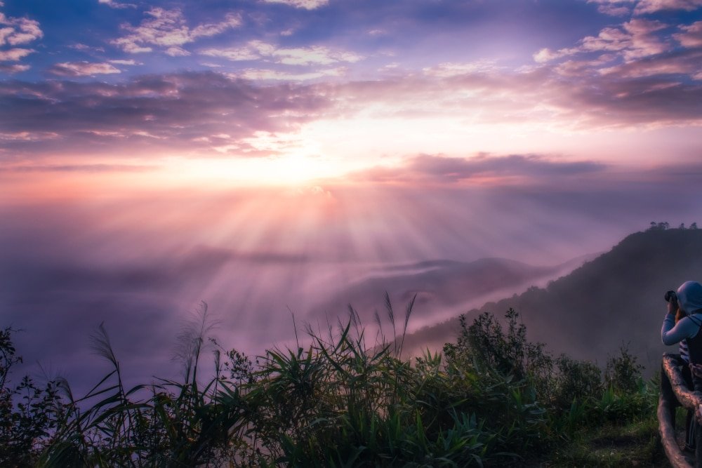 บุคคลหนึ่งถ่ายภาพหุบเขาที่ปกคลุมด้วยหมอกยามรุ่งอรุณ โดยมีแสงอาทิตย์ส่องผ่านเมฆและพืชพรรณ ที่เที่ยวปลายฝนต้นหนาว เขียวชอุ่มในเบื้องหน้า—ถ่ายทอดความมหัศจรรย์ของสถานที่ท่องเที่ยวปลายฝนต้นหนาวได้อย่างสมบูรณ์แบบ.