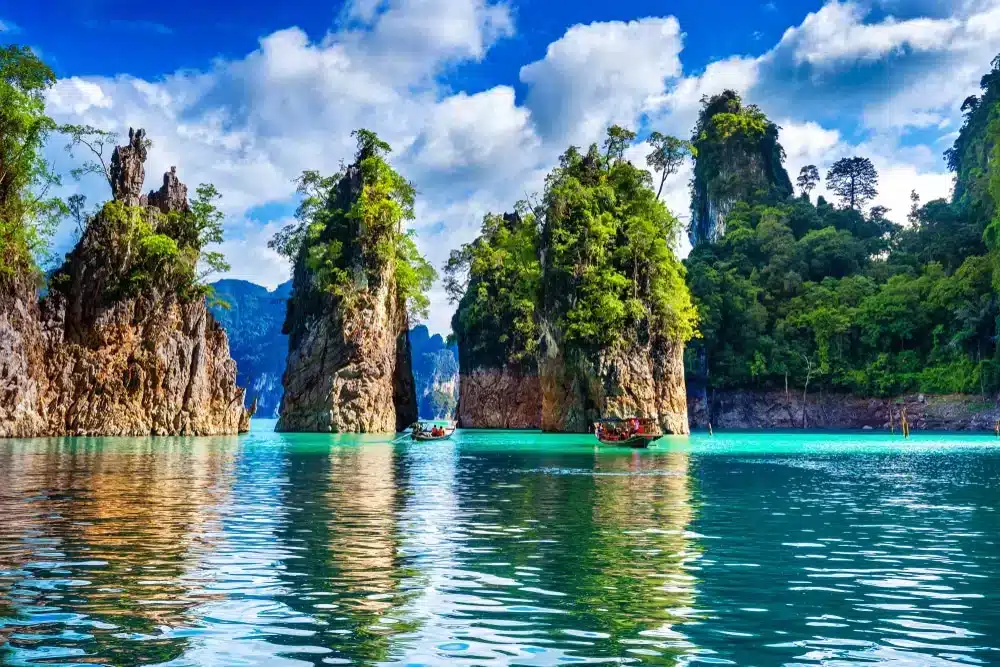 หน้าผาหินปูนสูงตระหง่านปกคลุมด้วยพืชพรรณสีเขียวขจี โอบล้อม เที่ยวสุราษฎร์ธานี ด้วยน้ำทะเลสีฟ้าเทอร์ควอยซ์ มีเรือลำเล็กลอยอยู่ใกล้เคียง—ภาพอันงดงามเหมาะสำหรับผู้ที่ต้องการสำรวจสถานที่ท่องเที่ยวในสุราษฎร์ธานีและค้นพบแหล่งท่องเที่ยวในสุราษฎร์ธานี.