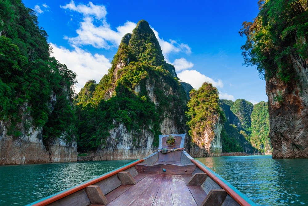 เรือไม้ลำหนึ่งแล่นไปบนผืนน้ำสีฟ้าอมเขียว ท่ามกลางหน้าผาหินปูนสูงตระหง่านปกคลุมด้วยป่าไม้ ทอดยาวใต้ท้องฟ้าสีครามแต้มด้วยเมฆขาวบางเบา—เป็นสถานที่ท่องเที่ยวทางธรรมชาติที่สมบูรณ์แบบสำหรับผู้รักธรรมชาติ.