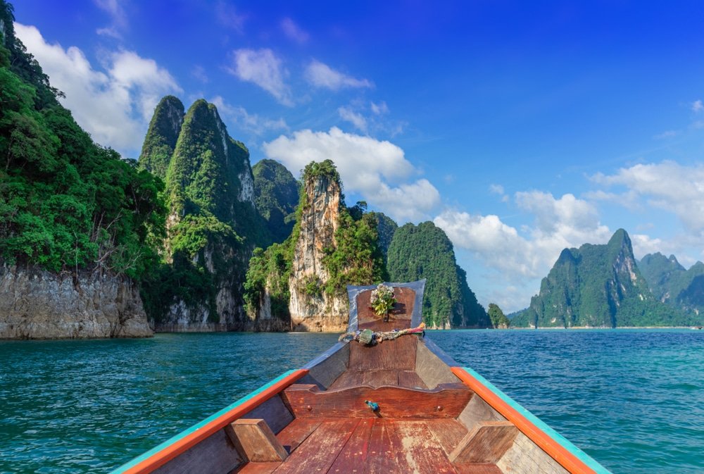 เรือไม้ลำหนึ่งมุ่งหน้าไปยังหน้าผาหินปูนและภูเขาเขียวขจีที่โผล่ขึ้นมา เที่ยวสุราษฎร์ธานี จากน้ำสีฟ้าภายใต้ท้องฟ้าแจ่มใส ถ่ายทอดความงดงามอันน่าทึ่งของสถานที่ท่องเที่ยวสุราษฎร์ธานี.