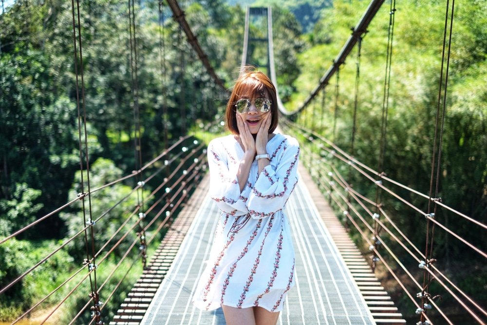 ผู้หญิงในชุดสีขาว ยืนอยู่บนสะพาน ที่เที่ยวสุราษฎร์ธานี แขวน ท่ามกลางธรรมชาติสีเขียวขจี กำลังกุมใบหน้าและมองกล้อง หนึ่งในสถานที่ท่องเที่ยวสุราษฎร์ธานี.
