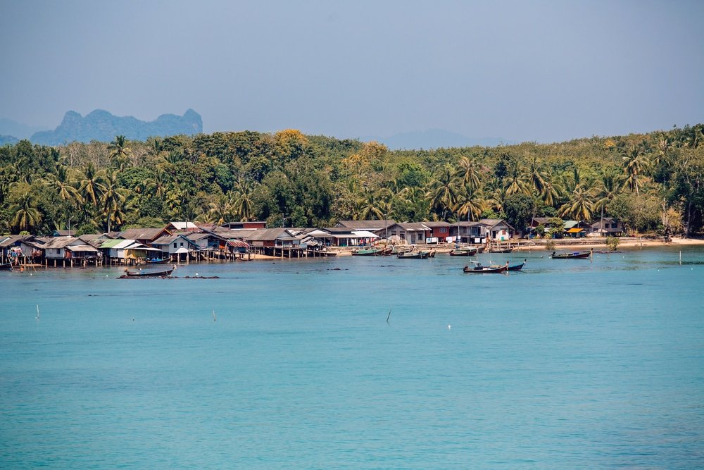 หมู่บ้านชายฝั่งที่มีบ้านไม้และเรือเรียงรายตามแนวชายฝั่ง ล้อมรอบด้วยต้นไม้และน้ำทะเลสีฟ้าใสภายใต้ท้องฟ้าแจ่มใส—เหมาะสำหรับผู้ที่มองหาสถานที่ท่องเที่ยวทางธรรมชาติ.