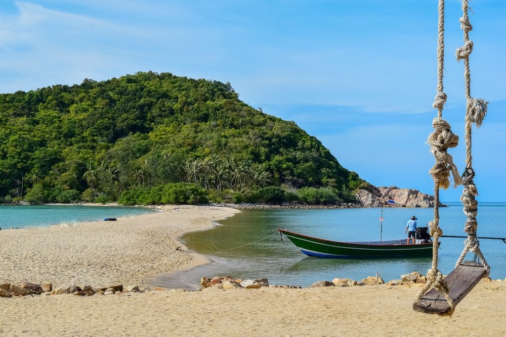 เรือไม้ลำหนึ่งจอดทอดสมออยู่ใกล้ชายฝั่งทราย โดยมีเนินเขาสีเขียว ที่เที่ยวสุราษฎร์ธานี เป็นฉากหลัง และมีชิงช้าเชือกแขวนอยู่จากต้นไม้ในเบื้องหน้า—ภาพทิวทัศน์อันงดงามของสุราษฎร์ธานี.