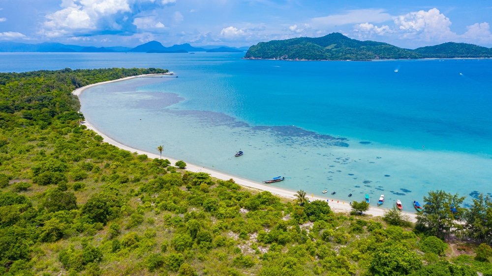 ภาพมุมสูงของชายหาดทรายโค้งในสุราษฎร์ธานีที่เที่ยว ล้อมรอบด้วยพืชพรรณสีเขียว น้ำทะเลใสสีฟ้าเท ที่เที่ยวสุราษฎร์ธานี อร์ควอยซ์ และภูเขาไกลโพ้นภายใต้ท้องฟ้าที่มีเมฆบางส่วน.