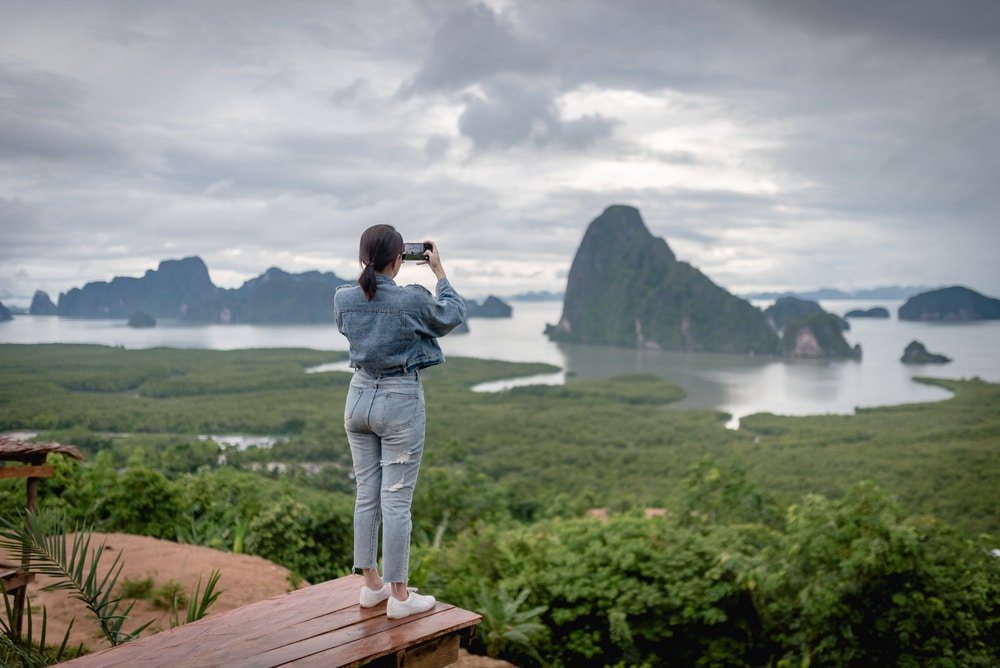บุคคลในชุดผ้าเดนิมยืนอยู่บนแพลตฟอร์มไม้ ณ สถานที่ท่องเที่ยวทางธรรมชาติ กำลังถ่ายรูปอ่าวที่สวยงามซึ่งมีเกาะสีเขียวและท้องฟ้าที่มีเมฆปกคลุม.