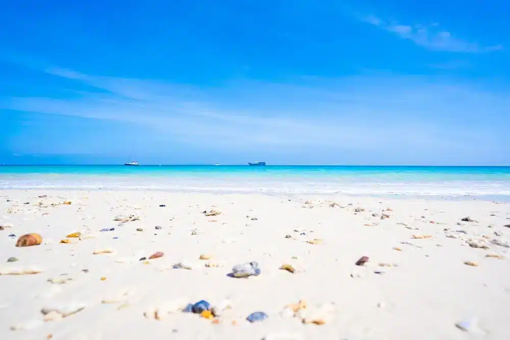 ชายหาดทรายที่มีหินเล็ก ๆ กระจัดกระจายอยู่เบื้องหน้า น้ำทะเลใสสีฟ้า และเรือสองลำลอยอยู่ไกลสุดขอบฟ้าภายใต้ท้องฟ้าสีฟ้าสดใส—เหมาะอย่างยิ่งสำหรับผู้ที่กำลังมองหสถานที่ท่องเที่ยวทางธรรมชาติ.