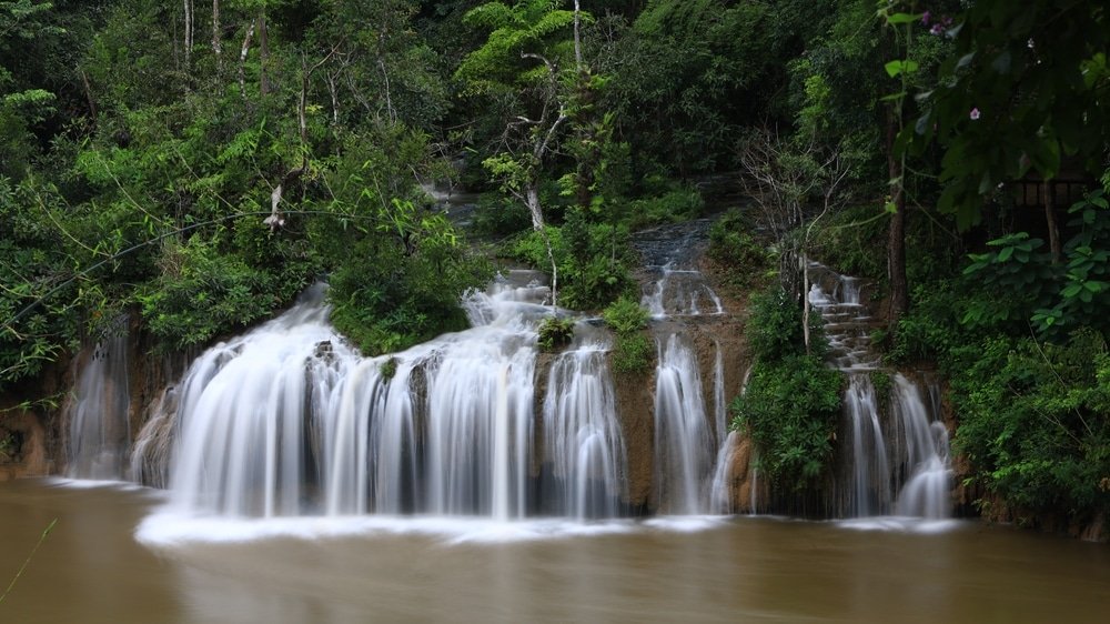น้ำตกไหลผ่านโขดหินที่ล้อมรอบด้วยพืชพรรณเขียวชอุ่ม น้ำตก ที่เที่ยวปลายฝนต้นหนาว กระทบลงสู่แม่น้ำที่มีโคลนเบื้องล่าง—สถานที่ที่เหมาะสำหรับคนรักธรรมชาติที่กำลังมองหา ที่เที่ยวปลายฝนต้นหนาว.