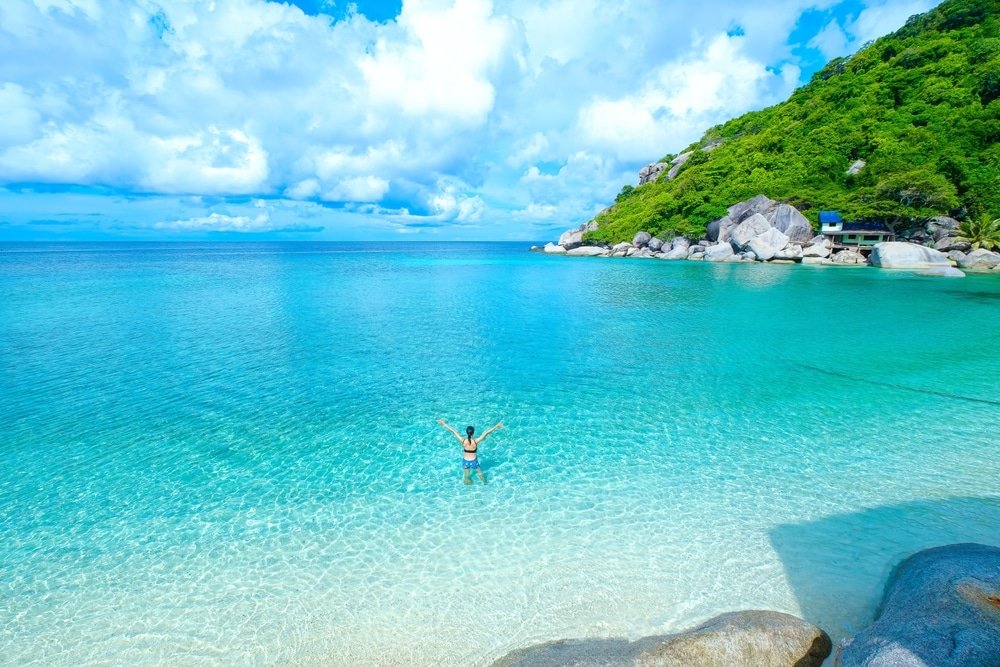 บุคคลหนึ่งยืนอยู่ในน้ำสีฟ้าใสตื้นใกล้ชายหาดเขตร้อนที่มีเนินเขาสีเขียวและหินเป็นฉากหลัง เป็นภาพ ที่เที่ยวสุราษฎร์ธานี ที่สมบูรณ์แบบสำหรับผู้ที่ต้องการสำรวจสถานที่ท่องเที่ยวสุราษฎร์ธานีภายใต้ท้องฟ้าที่มีเมฆบางส่วน.