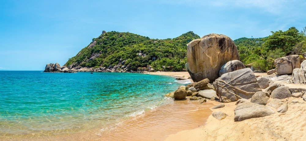 ชายหาดทรายขาวสะอาด น้ำทะเลสีฟ้าใสราวกับเทอร์ควอยซ์ หาดหินใหญ่ และภูเขาเขียวชอุ่มภายใต้ท้องฟ้าสีครามสดใส—เหมาะสำหรับผู้ที่รักการท่องเที่ยวธรรมชาติ.
