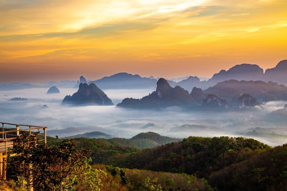 ทิวทัศน์ภูเขาในยามพระอาทิตย์ขึ้น พร้อมชั้นหมอกที่โอบล้อมเนินเขา ท้องฟ้าสีสันสวยงาม และจุดชมวิวไม้ในเบื้องหน้า—เหมาะสำหรับผู้ที่มองหาสถานที่ท่องเที่ยวทางธรรมชาติและบรรยากาศผ่อนคลาย.
