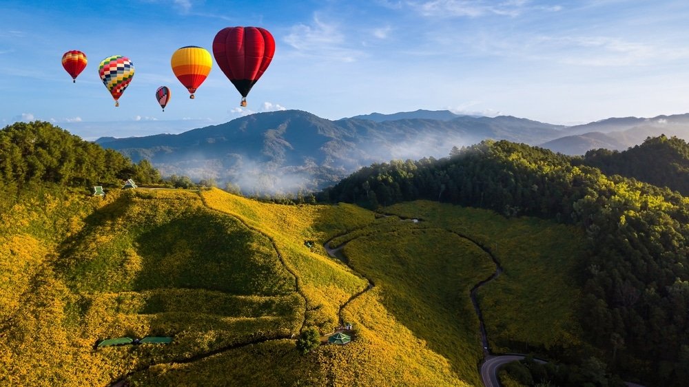 บอลลูนลมร้อนหลากสีลอยอยู่เหนือเนินเขาที่ปกคลุมไปด้วยดอกไม้สีเหลือง—ภาพที่งดงามราวกับฝันที่ทำให้ที่นี่กลายเป็นสถานที่ท่องเที่ยวที่เหมาะอย่างยิ่งสำหรับปลายฝนต้นหนาว พร้อมถนนที่คดเคี้ยวและภูเขาที่งดงามภายใต้ท้องฟ้าสีครามใส.