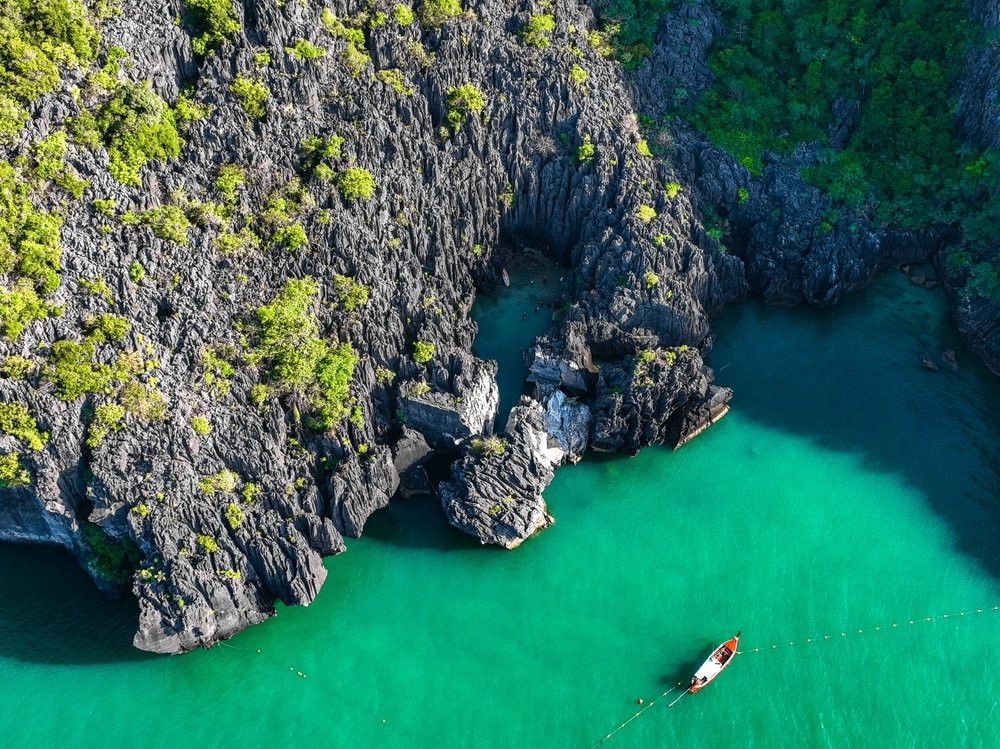 ภาพมุมสูงของเรือลำเล็กที่ลอยอยู่ใกล้หน้าผาหินปูนและน้ำทะเลสีฟ้าใสราวคริสตัล พร้อมด้วยพื้นที่สีเขียวบนชายฝั่งหิน—เหมาะสำหรับผู้ที่มองหาสถานที่ท่องเที่ยวทางธรรมชาติที่น่าประทับใจ.