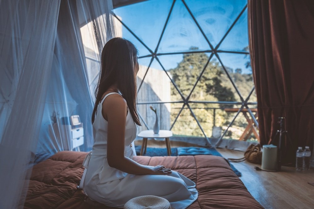 ผู้หญิงในชุดสีขาว นั่งอยู่บนเตียงในห้องทรงโดม มองออกไปผ่าน ที่เที่ยวปลายฝนต้นหนาว หน้าต่างรูปทรงเรขาคณิตขนาดใหญ่ไปยังต้นไม้ภายนอก—ช่วงเวลาที่สงบเงียบเหมาะสำหรับการท่องเที่ยวในช่วงปลายฝนต้นหนาว.