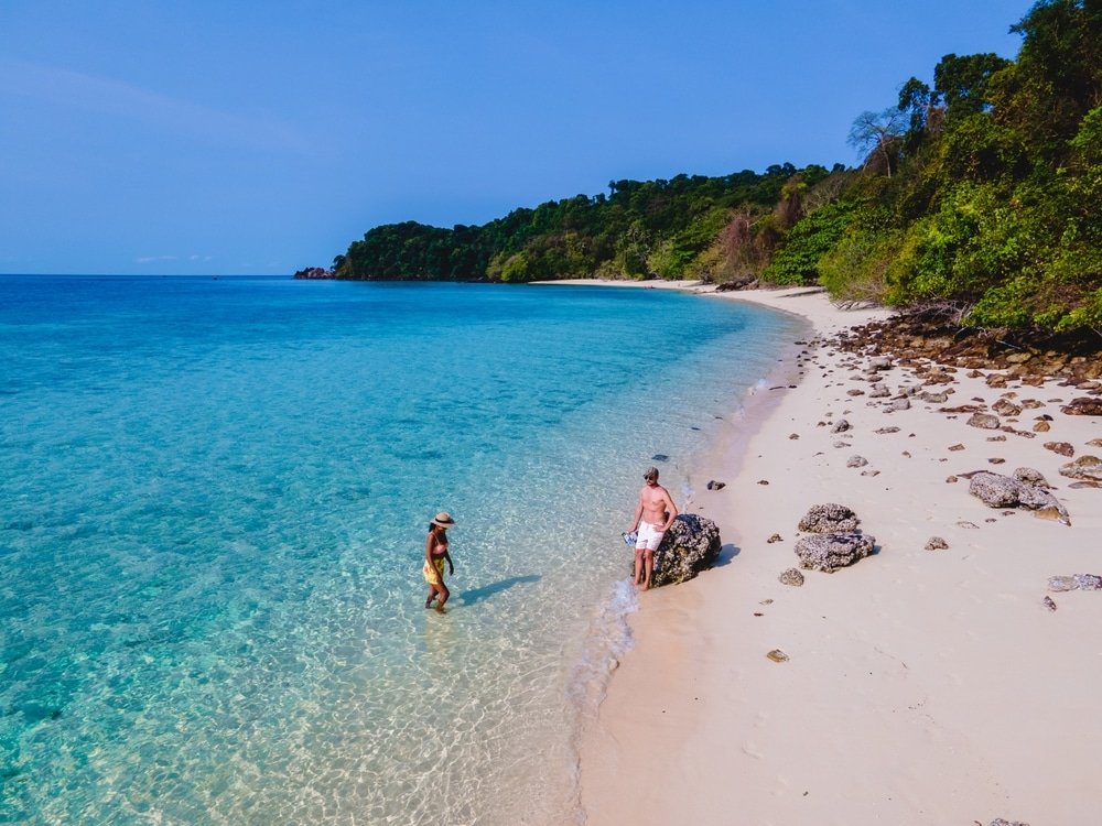 สองคนยืนอยู่บนชายหาดทรายข้างน้ำทะเลใสสีฟ้า ล้อมรอบด้วยพื้นที่หินและต้นไม้เขียวชอุ่มหนาแน่น—เหมาะสำหรับคนรักธรรมชาติ—ภายใต้ท้องฟ้าแจ่มใส.
