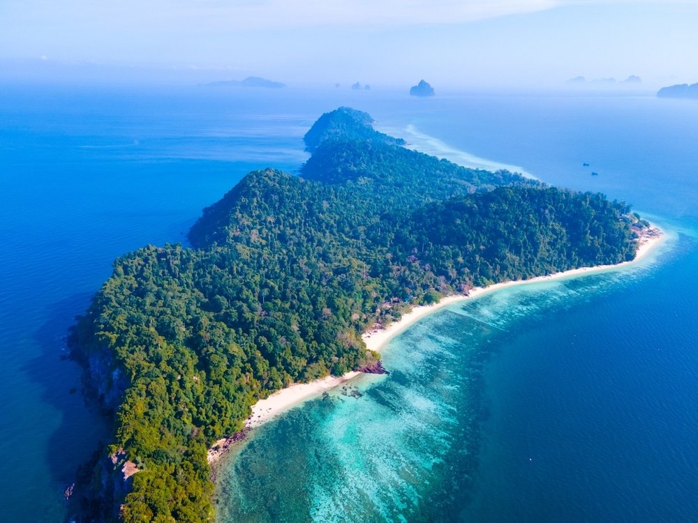 ภาพมุมสูงของเกาะที่เขียวชอุ่ม—หนึ่งในสถานที่ท่องเที่ยวทางธรรมชาติที่ดีที่สุด—พร้อมชายหาดทรายและน้ำทะเลสีฟ้าใสล้อมรอบด้วยมหาสมุทรภายใต้ท้องฟ้าสีคราม.