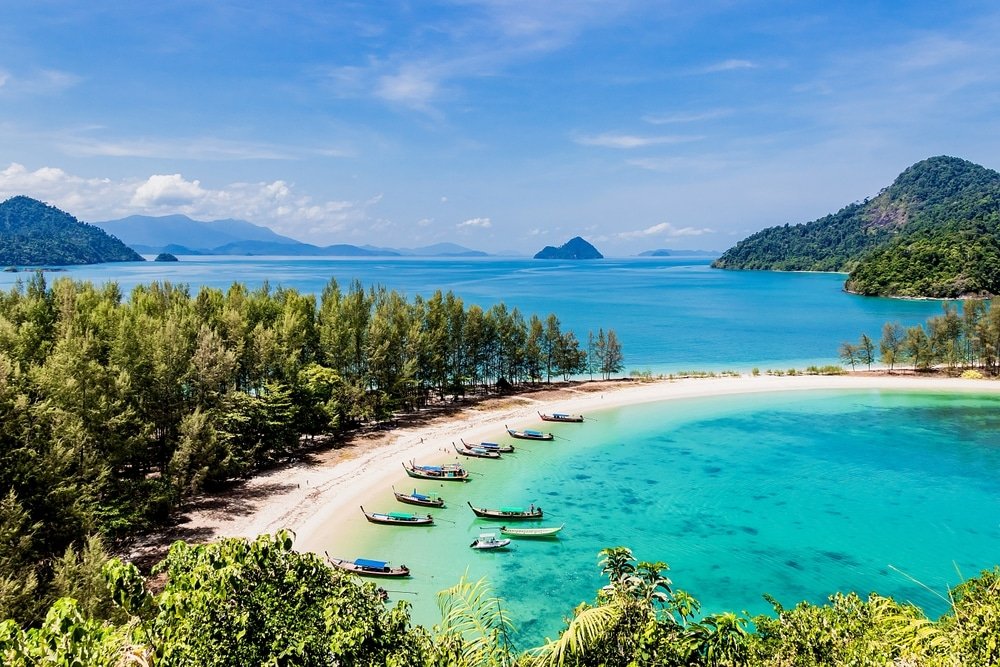 ชายหาดทรายโค้งน้ำสีฟ้าคราม มีเรือหลายลำจอดอยู่ใกล้ชายฝั่ง และภูเขาเขียวขจีเป็นฉากหลังภายใต้ท้องฟ้าสีครามใส—เหมาะสำหรับผู้ที่มองหาสถานที่ท่องเที่ยวทางธรรมชาติ.