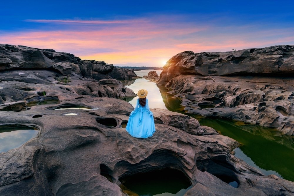 บุคคลในชุดเดรสและหมวกสีน้ำเงิน ที่เที่ยวปลายฝนต้นหนาว ยืนอยู่บนพื้นหินใกล้แม่น้ำในยามพระอาทิตย์ตกดิน ล้อมรอบด้วยหินขนาดใหญ่และแอ่งน้ำ—เหมาะสำหรับผู้ที่รักการเดินทางมองหาสถานที่ท่องเที่ยวในช่วงปลายฝนต้นหนาว.