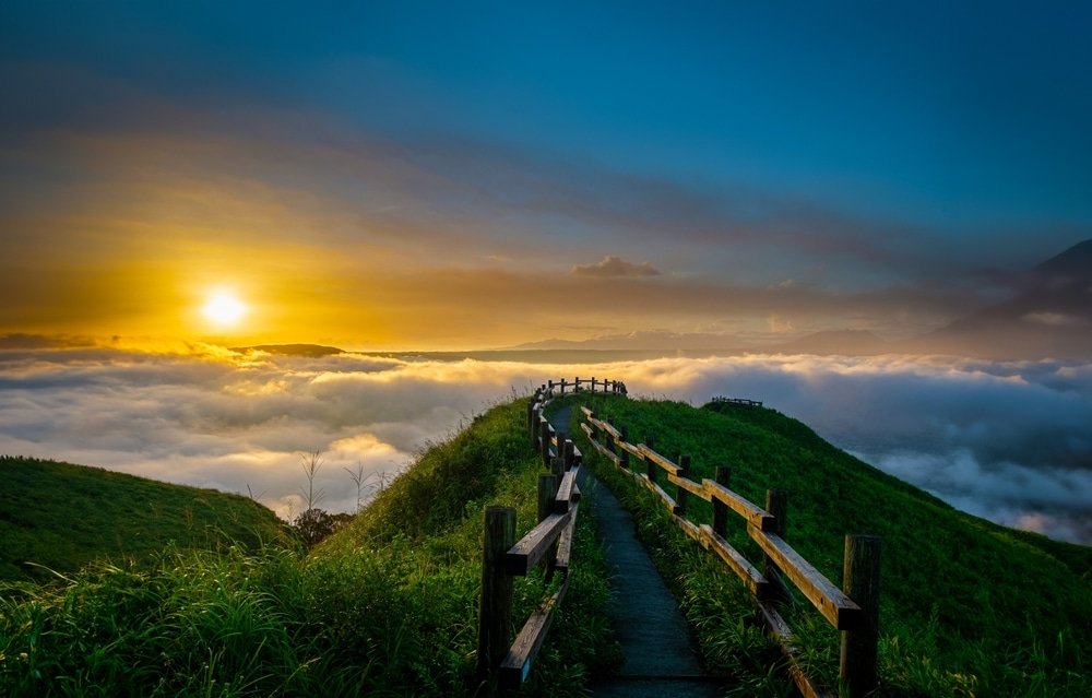 เส้นทางแคบปูด้วยหินที่มีรั้วไม้คดเคี้ยวไปตามยอดเขาที่ปกคลุมด้วย ที่เที่ยวปลายฝนต้นหนาว หญ้าเหนือเมฆยามพระอาทิตย์ขึ้น—สถานที่ท่องเที่ยวในฤดูฝนปลายฤดูฝนต้นฤดูหนาวที่งดงาม—กับดวงอาทิตย์ต่ำบนขอบฟ้าและภูเขาในระยะไกล.