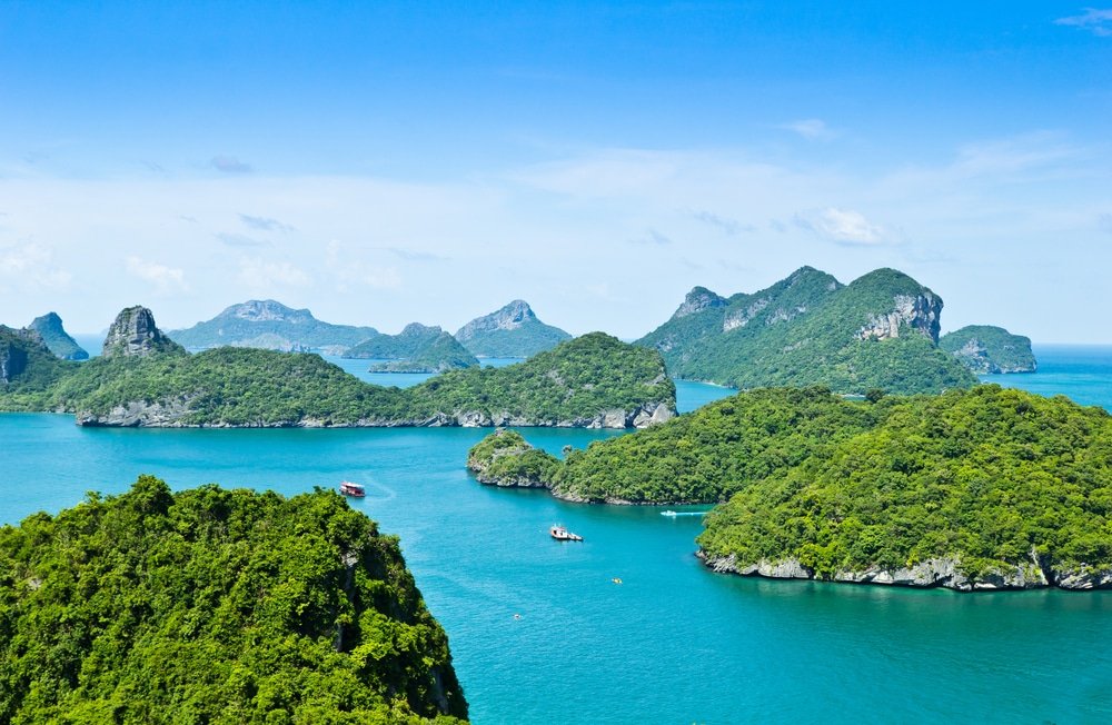 ภาพมุมสูงของเกาะเขียวชอุ่มที่รายล้อมไปด้วยน้ำทะเลสีฟ้าสดใสภาย ที่เที่ยวสุราษฎร์ธานี ใต้ท้องฟ้าที่แจ่มใส สามารถมองเห็นเรือหลายลำที่แล่นอยู่ แสดงให้เห็นถึงความงดงามของสุราษฎร์ธานีที่เที่ยว สถานที่ท่องเที่ยวที่คุณไม่ควรพลาดสำหรับการผจญภัยครั้งต่อไปของคุณ.