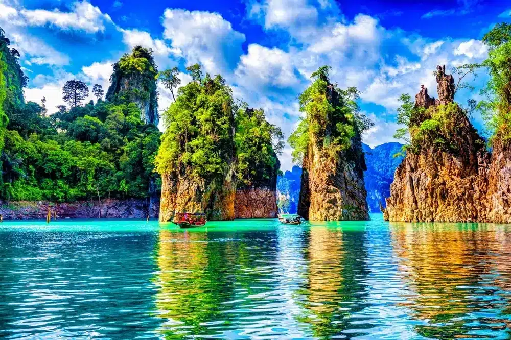 หินปูนสูงตระหง่านปกคลุมด้วยพืชพรรณสีเขียวโผล่ขึ้นจากน้ำสีฟ้าใส ที่เที่ยวปลายฝนต้นหนาว ราวคริสตัล มีเรือลำเล็กลอยอยู่ใกล้ๆ ภายใต้ท้องฟ้าที่มีเมฆบางส่วน—สถานที่ท่องเที่ยวในฤดูฝนปลายฤดูฝนต้นฤดูหนาวที่เหมาะสำหรับผู้รักธรรมชาติที่ต้องการชมทัศนียภาพที่สวยงามตระการตา.