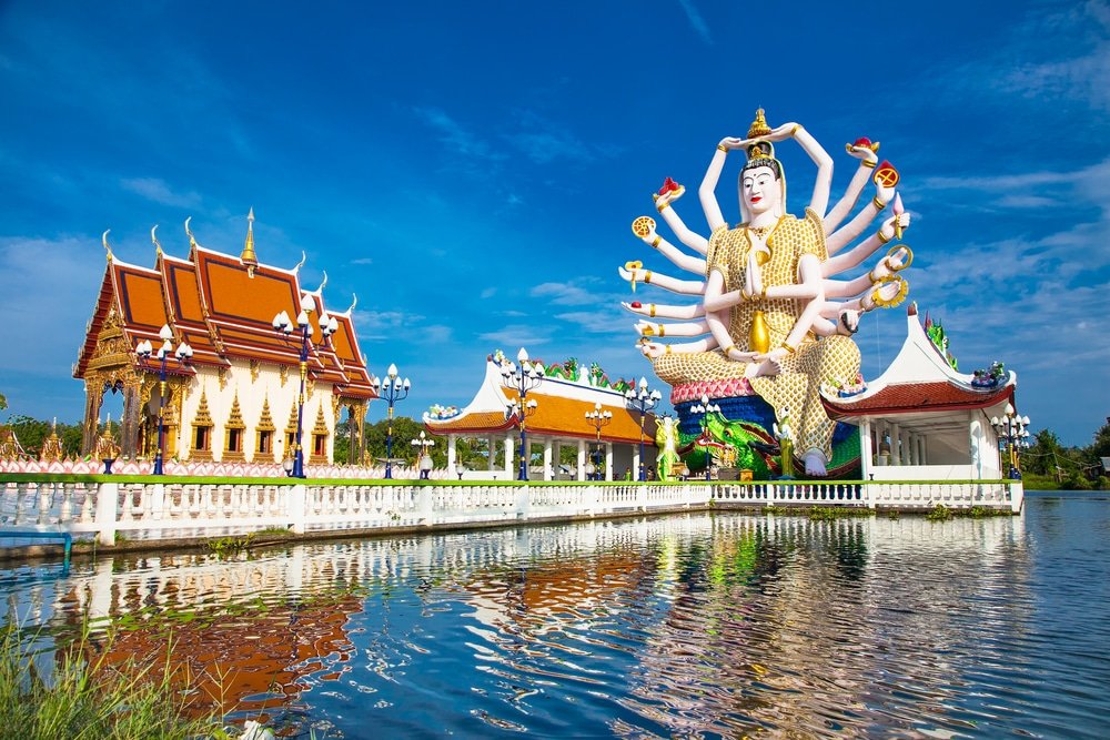 รูปปั้นขนาดใหญ่หลายแขนและ ที่เที่ยวสุราษฎร์ธานี อาคารวัดไทยแบบดั้งเดิมตั้งอยู่ริมสระน้ำใต้ท้องฟ้าสีครามที่วัดพระใหญ่ เกาะสมุย หนึ่งในสถานที่ท่องเที่ยวสุราษฎร์ธานีที่ต้องไปเยือนสำหรับผู้ที่ต้องการสำรวจเที่ยวสุราษฎร์ธานี.