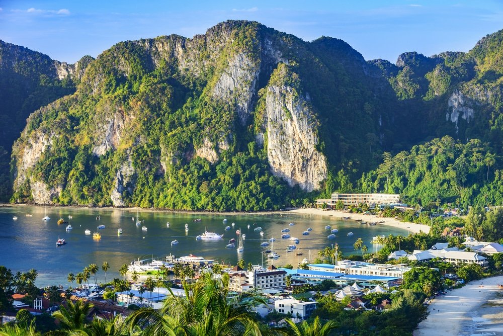 ทัศนียภาพของอ่าวที่มีเรือจอดทอดสมออยู่ในน้ำ ล้อมรอบด้วยหน้าผาสีเขียวและเมืองเล็กๆ ตามแนวชายฝั่งใต้ท้องฟ้าที่แจ่มใส—สถานที่ท่องเที่ยวทางธรรมชาติที่สมบูรณ์แบบสำหรับการพักผ่อนและความงามของทิวทัศน์.