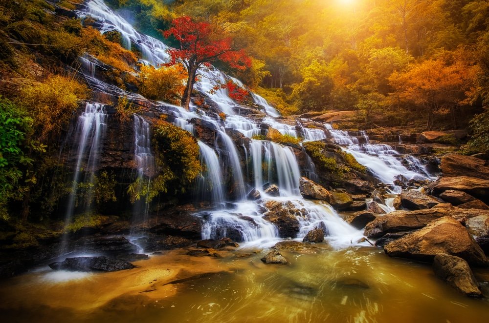 น้ำตกหลายชั้นไหลลดหลั่นลงมาบนโขดหินสู่แอ่งน้ำสงบ รายล้อมด้วย ที่เที่ยวปลายฝนต้นหนาว ใบไม้ฤดูใบไม้ร่วงที่เขียวขจีและแสงอาทิตย์ที่ส่องประกายเป็นฉากหลัง—สถานที่ท่องเที่ยวปลายฝนต้นหนาวที่สมบูรณ์แบบสำหรับคนรักธรรมชาติ.