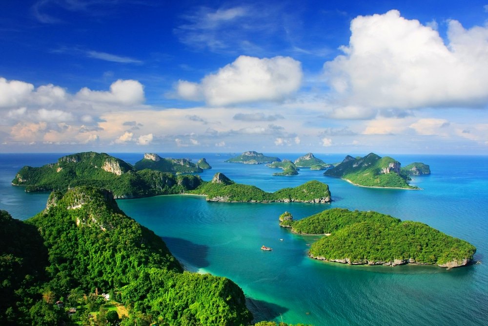 ภาพมุมสูง ที่เที่ยวสุราษฎร์ธานี ของเกาะสีเขียวที่ปกคลุมไปด้วยป่าไม้ ล้อมรอบด้วยทะเลสีฟ้าเทอร์ควอยซ์ภายใต้ท้องฟ้าที่มีเมฆบางส่วน ถ่ายทอดความงดงามตามธรรมชาติของสถานที่ท่องเที่ยวสุราษฎร์ธานี สำหรับผู้ที่ต้องการเที่ยวสุราษฎร์ธานี.