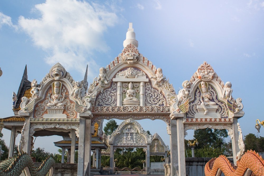 ประตูวัดสีขาวประดับประดาอย่าง ที่เที่ยวสุราษฎร์ธานี วิจิตร พร้อมด้วยงานแกะสลักและรูปปั้นที่ประณีต ตั้งตระหง่านท่ามกลางท้องฟ้าสีครามและธรรมชาติเขียวขจี—สถานที่ท่องเที่ยวสุราษฎร์ธานีที่ไม่ควรพลาดสำหรับผู้ที่ต้องการสัมผัสความงดงามของแหล่งท่องเที่ยวสุราษฎร์ธานี.
