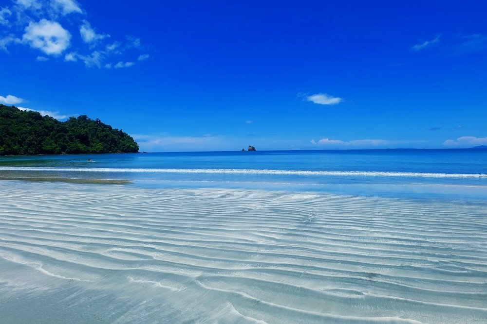 ชายหาดทรายที่มีลวดลายเป็นคลื่น น้ำทะเลใสสีฟ้า ภูเขาเขียวขจีในระยะไกล และท้องฟ้าสีฟ้าสดใสที่มีเมฆกระจายอยู่—เหมาะสำหรับผู้ที่มองหาสถานที่ท่องเที่ยวทางธรรมชาติ.