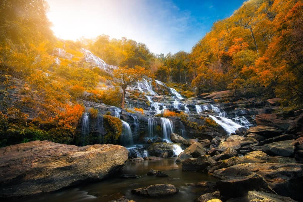 น้ำตกหลายชั้นไหลผ่านโขดหินที่ล้อมรอบด้วยต้นไม้ใบไม้สีส้มในฤดูใบไม้ร่วง ภายใต้แสงแดดจ้าและท้องฟ้าสีคราม—เหมาะอย่างยิ่ง ที่เที่ยวปลายฝนต้นหนาว สำหรับการพักผ่อนในช่วงปลายฝนต้นหนาว.