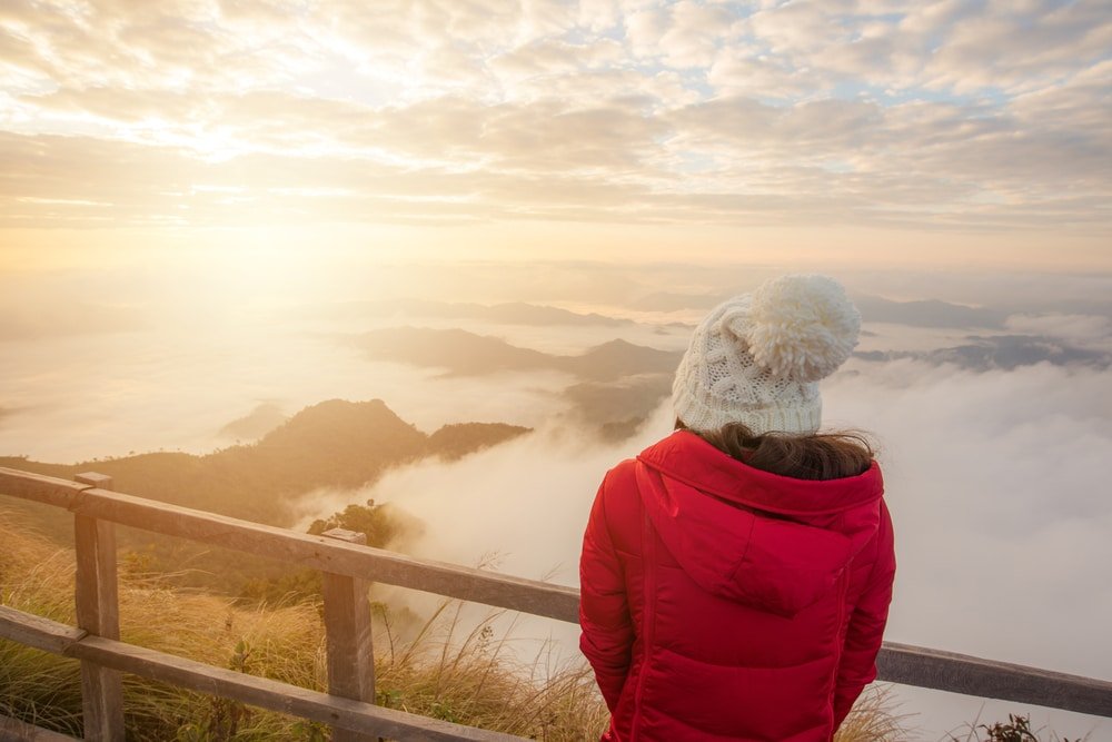 บุคคลในเสื้อแจ็คเก็ตสีแดงและหมวกถักสีขาว ยืนอยู่ที่ราวไม้ มองดู ที่เที่ยวปลายฝนต้นหนาว หุบเขาที่มีเมฆปกคลุมและภูเขาในยามพระอาทิตย์ขึ้นหรือพระอาทิตย์ตก—เหมาะสำหรับคนรักการท่องเที่ยวในช่วงปลายฝนต้นหนาว.