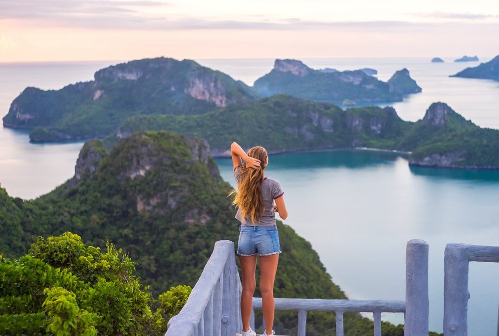 คนผมยาวคนหนึ่งยืนอยู่บนจุดชมวิวที่มองเห็นสถานที่ท่องเที่ยวทางธรรมชาติ มีเกาะสีเขียวและน้ำทะเลสีฟ้าสงบในยามพระอาทิตย์ตก.