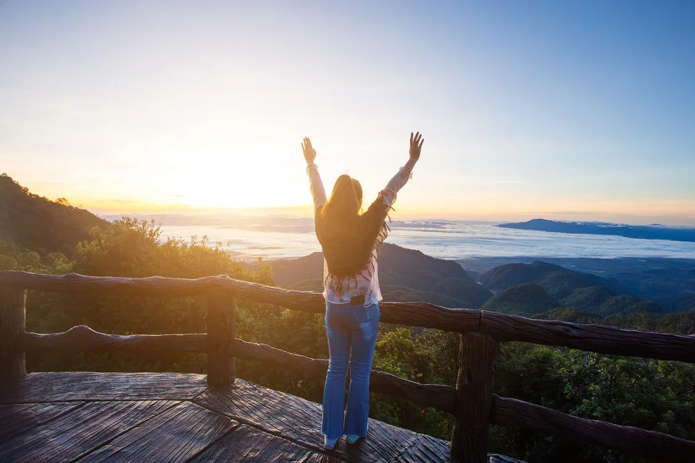 บุคคลหนึ่งยืนอยู่บนดาดฟ้าไม้ หันหน้าไปทางภูเขาและพระอาทิตย์ขึ้น ที่เที่ยวปลายฝนต้นหนาว พร้อมยกแขนขึ้น—ดื่มด่ำกับความสงบของสถานที่ท่องเที่ยวปลายฝนต้นหนาวใต้ท้องฟ้าที่แจ่มใสและเมฆที่ปกคลุมหุบเขาเบื้องล่าง.