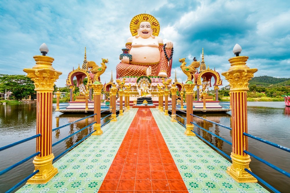 พระพุทธรูปขนาดใหญ่ประทับนั่งล้อมรอบด้วยเสาและโครงสร้าง ที่เที่ยวสุราษฎร์ธานี ประดับประดาอย่างวิจิตร ตั้งอยู่บนแท่นเหนือผืนน้ำ โดยมีภูเขาและเมฆเป็นฉากหลัง—เป็นจุดอันเป็นสัญลักษณ์สำหรับการท่องเที่ยวสุราษฎร์ธานี.