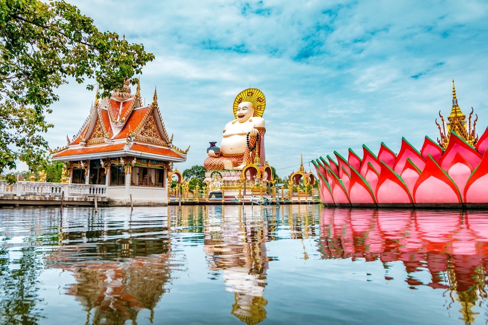 พระพุทธรูปขนาดใหญ่ วัดไทยแบบดั้งเดิม และโครงสร้างรูปดอกบัวสีชมพูสะท้อนอยู่ในน้ำสงบใต้ท้องฟ้าสีคราม ที่เที่ยวสุราษฎร์ธานี ณ หนึ่งในสถานที่ท่องเที่ยวที่ต้องไปเยือนในสุราษฎร์ธานี.