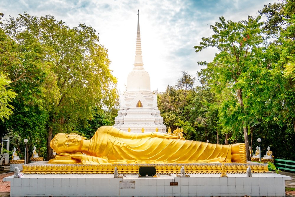 พระพุทธไสยาสน์ทองคำขนาดใหญ่ ที่เที่ยวสุราษฎร์ธานี ประดิษฐานอยู่เบื้องหน้าเจดีย์สีขาว รายล้อมไปด้วยต้นไม้เขียวขจีและท้องฟ้าสดใส—เป็นหนึ่งในสถานที่ท่องเที่ยวสุราษฎร์ธานีที่ไม่ควรพลาดสำหรับผู้ที่วางแผนจะเที่ยวสุราษฎร์ธานี.