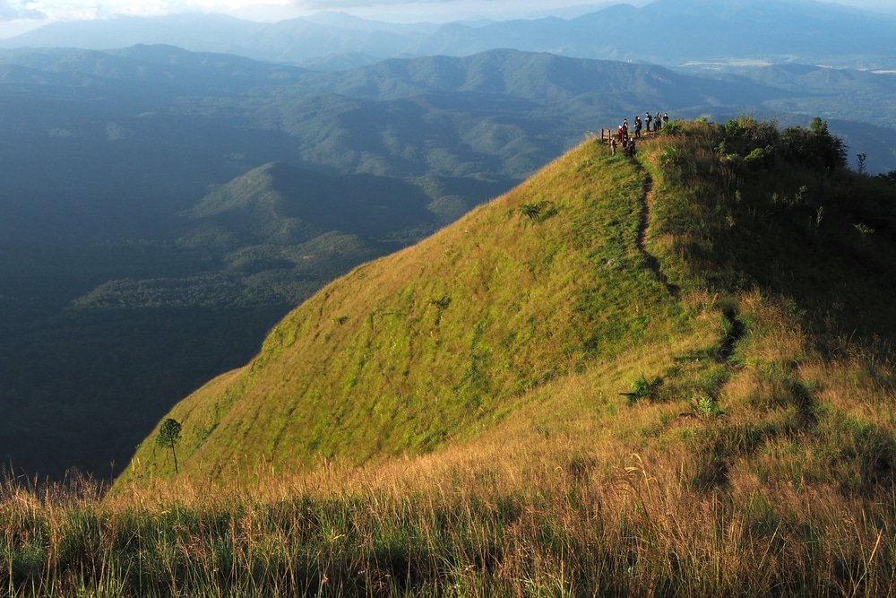 กลุ่มคนยืนอยู่บนยอดเขาที่ปกคลุมด้วยหญ้า มีเส้นทางแคบ ๆ ที่ ที่เที่ยวปลายฝนต้นหนาว สามารถมองเห็นได้ทอดยาวออกไป เห็นภูเขาและหุบเขาอยู่เบื้องล่าง—สถานที่ท่องเที่ยวที่เหมาะอย่างยิ่งสำหรับผู้คนที่ต้องการชมวิวที่สวยงามอย่างน่าทึ่งในช่วงปลายฝนต้นหนาว.