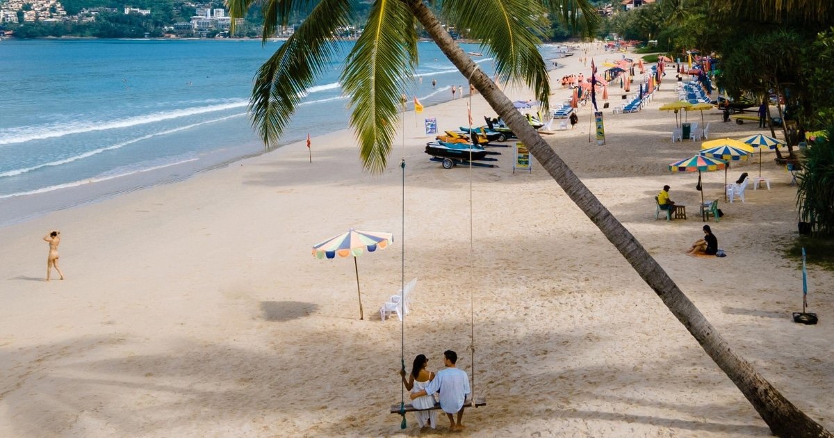 คู่รักนั่งอยู่บนชิงช้าใต้ต้นปาล์มบนชายหาดทราย กำลังเพลิดเพลินกับสถานที่ท่องเที่ยวธรรมชาติ พร้อมร่มและผู้คนในฉากหลังใกล้ชายฝั่ง.
