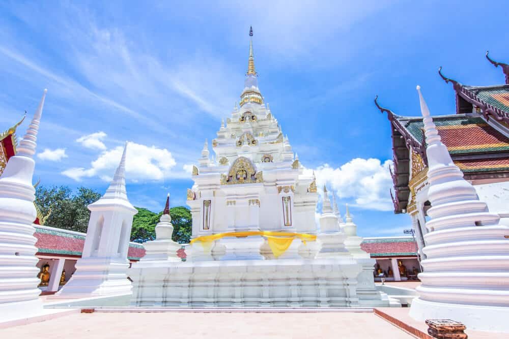 เจดีย์พุทธสีขาวประดับด้วยราย ที่เที่ยวสุราษฎร์ธานี ละเอียดสีทอง ตั้งอยู่ใต้ท้องฟ้าสีฟ้าสดใส ล้อมรอบด้วยเจดีย์ขนาดเล็กและอาคารวัด ทำให้เป็นสถานที่ท่องเที่ยวที่ต้องไปเยือนในสุราษฎร์ธานี สำหรับผู้ที่ต้องการเที่ยวสุราษฎร์ธานี.