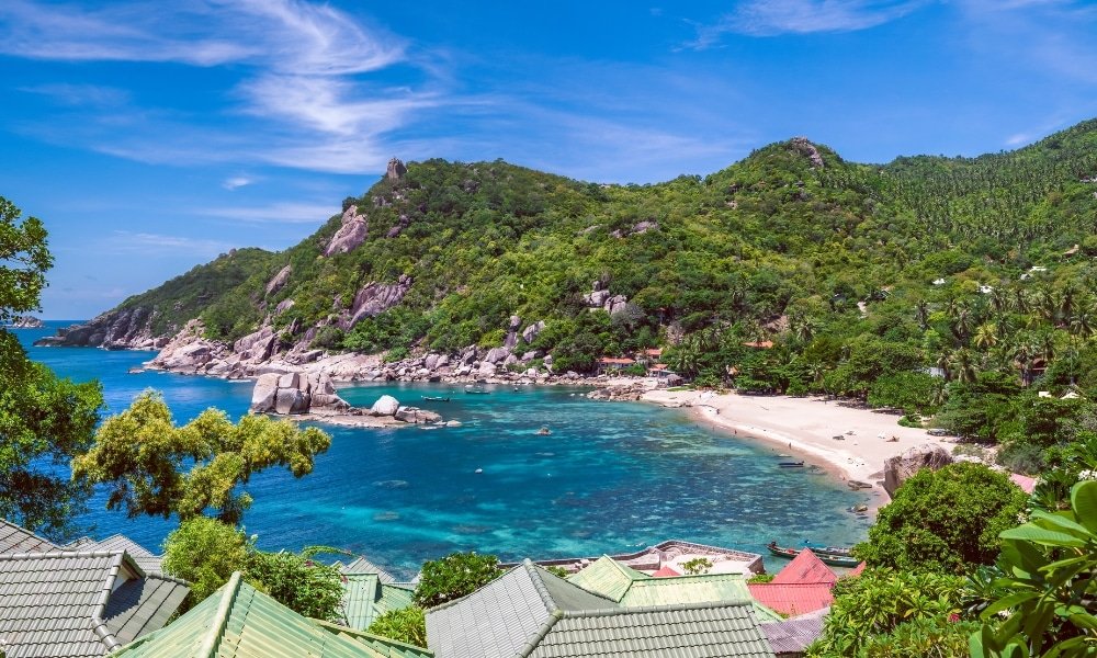 ชายหาดเขตร้อนที่มีน้ำทะเลสีเทอร์ควอยซ์ ทรายขาว ภูเขาเขียว และหลังคาบ้านในเบื้องหน้า ท่ามกลางท้องฟ้าสีคราม—เหมาะสำหรับผู้ที่ชื่นชอบการท่องเที่ยวธรรมชาติ.