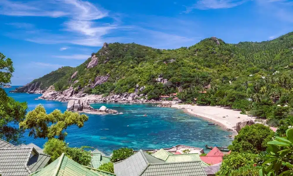 อ่าวเขตร้อนที่มีน้ำทะเลใสสีฟ้า ที่เที่ยวสุราษฎร์ธานี ชายหาดทรายขาว ภูเขาเขียวขจี และหลังคาบ้านหลายหลังในเบื้องหน้า ภายใต้ท้องฟ้าสดใสและแสงแดดจ้า—เหมาะอย่างยิ่งสำหรับผู้ที่ต้องการพักผ่อนและสำรวจความงามตามธรรมชาติใน สุราษฎร์ธานีที่เที่ยว.