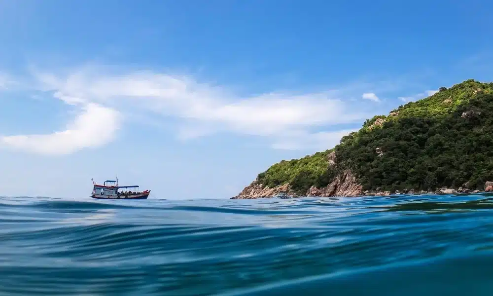 เรือลำเล็กกำลังลอยอยู่บนน้ำสีฟ้า ที่เที่ยวสุราษฎร์ธานี สงบใกล้กับเนินเขาที่ปกคลุมไปด้วยต้นไม้สีเขียวภายใต้ท้องฟ้าที่แจ่มใส—ภาพที่น่าดึงดูดใจเหมาะสำหรับผู้ที่กำลังมองหาสถานที่ท่องเที่ยวในสุราษฎร์ธานีที่เงียบสงบและสวยงาม.
