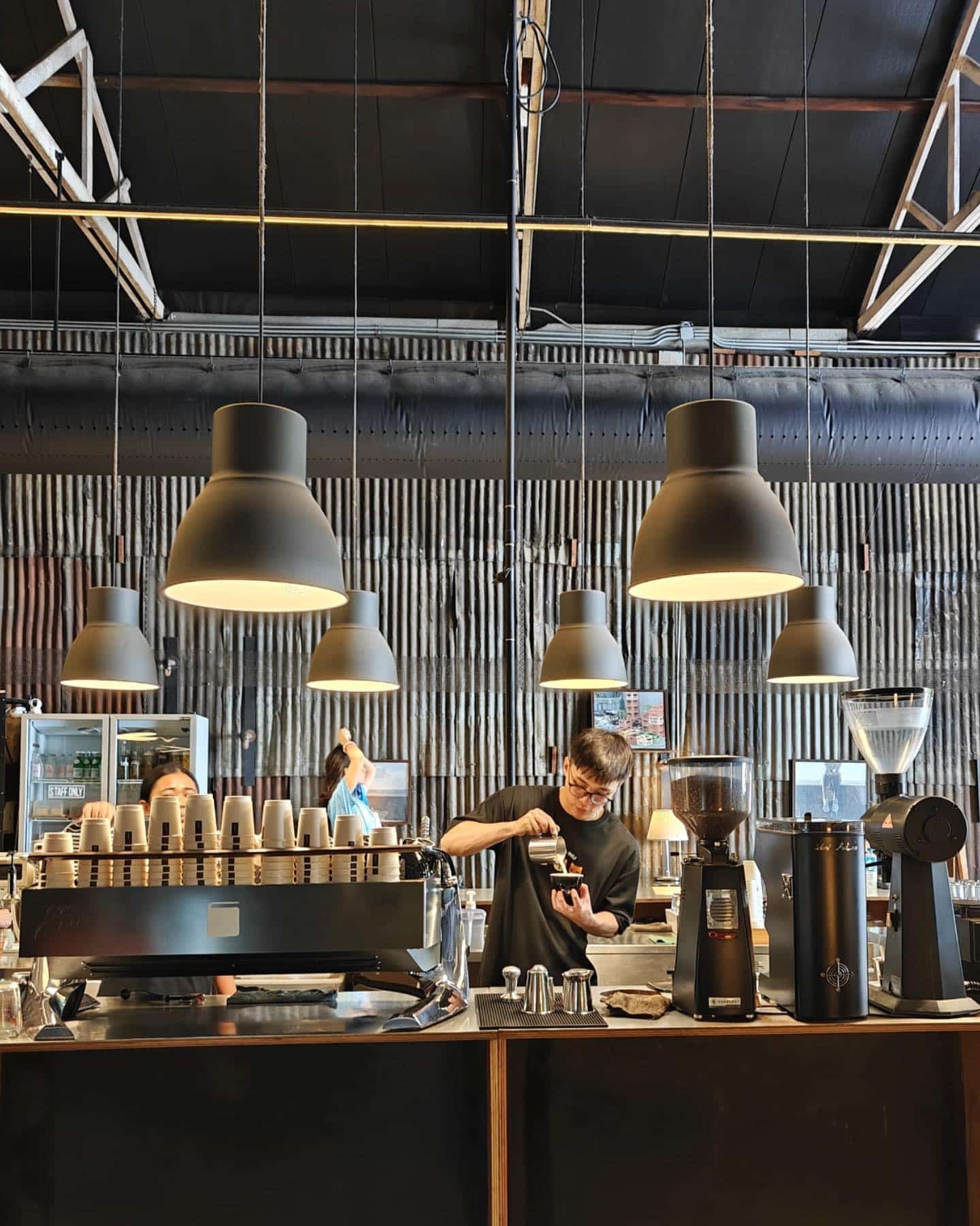 บาริสต้าเตรียมกาแฟอยู่หลัง ที่เที่ยวกรุงเทพ เคาน์เตอร์ของคาเฟ่สไตล์อุตสาหกรรม ซึ่งเป็นหนึ่งในสถานที่ท่องเที่ยวที่ซ่อนตัวอยู่ในกรุงเทพฯ พร้อมไฟแขวนและผนังโลหะลูกฟูกเป็นฉากหลัง.