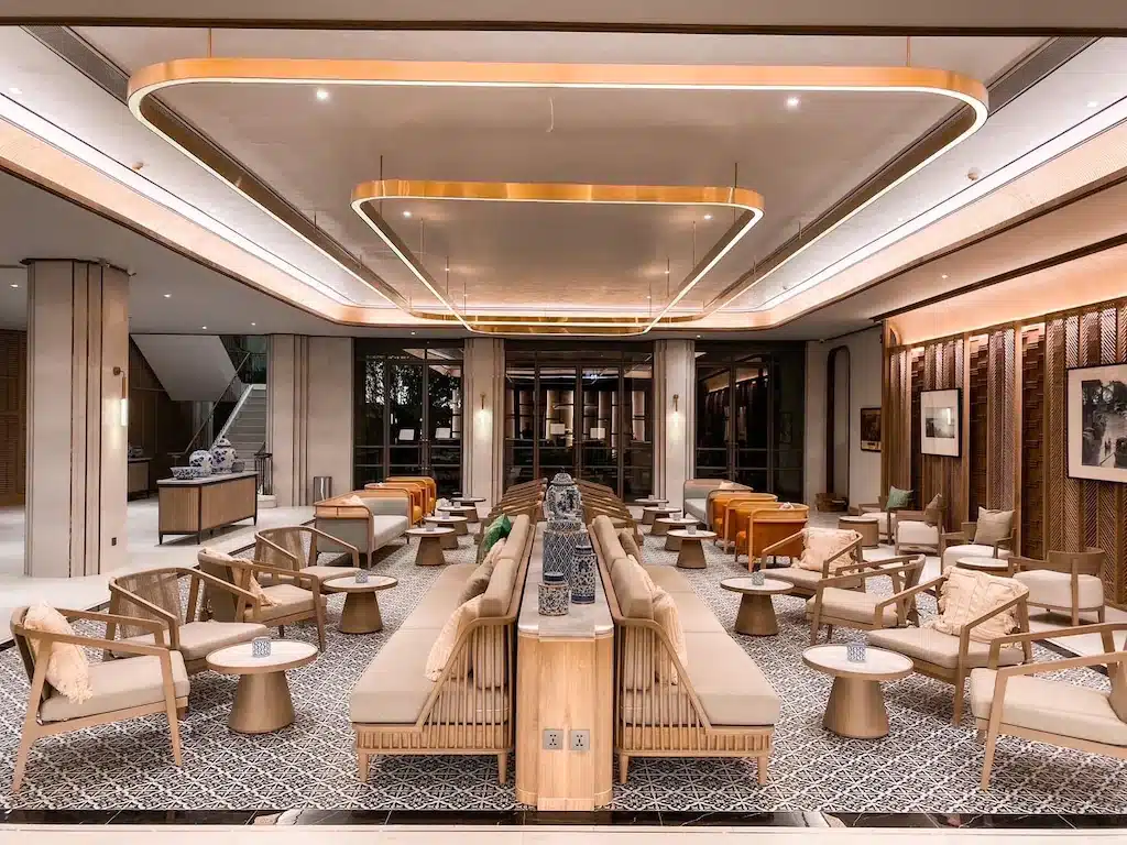 ล็อบบี้โรงแรมสมัยใหม่, ที่พักสุดชิคสำหรับนักท่องเที่ยวที่กำลังมอง ที่เที่ยวกรุงเทพ หาสถานที่ท่องเที่ยวในกรุงเทพฯ, โดดเด่นด้วยที่นั่งไม้แบบสมมาตร, หมอนอิงสีเบจ, โต๊ะกลมขนาดเล็ก, พื้นกระเบื้องลวดลาย, และแสงไฟเหนือศีรษะที่อบอุ่น.