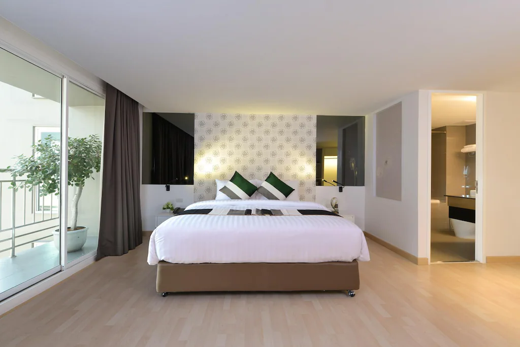 ห้องพักโรงแรมสมัยใหม่ใกล้แหล่งท่องเที่ยวในกรุงเทพมหานคร  ที่เที่ยวกรุงเทพ ประกอบด้วยเตียงขนาดใหญ่พร้อมผ้าปูที่นอนสีขาว หมอนตกแต่งสีเขียว ผนังตกแต่งลวดลาย พื้นไม้ ระเบียง และห้องน้ำแบบเปิดโล่ง.