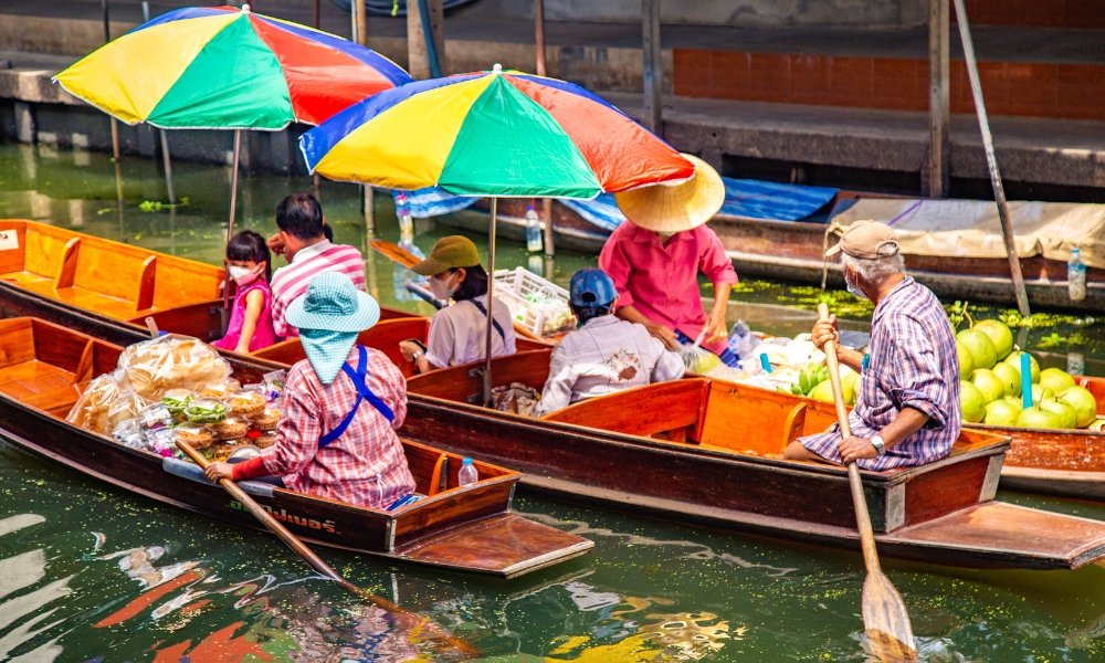 พ่อค้าแม่ค้าในเรือไม้ขายผักผลไม้และสินค้าต่างๆ ใต้ร่มสีสันสดใสที่ตลาดน้ำคึกคักบนคลอง ทำให้ที่นี่เป็นสถานที่ท่องเที่ยวที่ต้องไป ที่เที่ยวกรุงเทพ เยือนในกรุงเทพฯ สำหรับทุกคนที่มาสำรวจเมืองที่มีชีวิตชีวา.