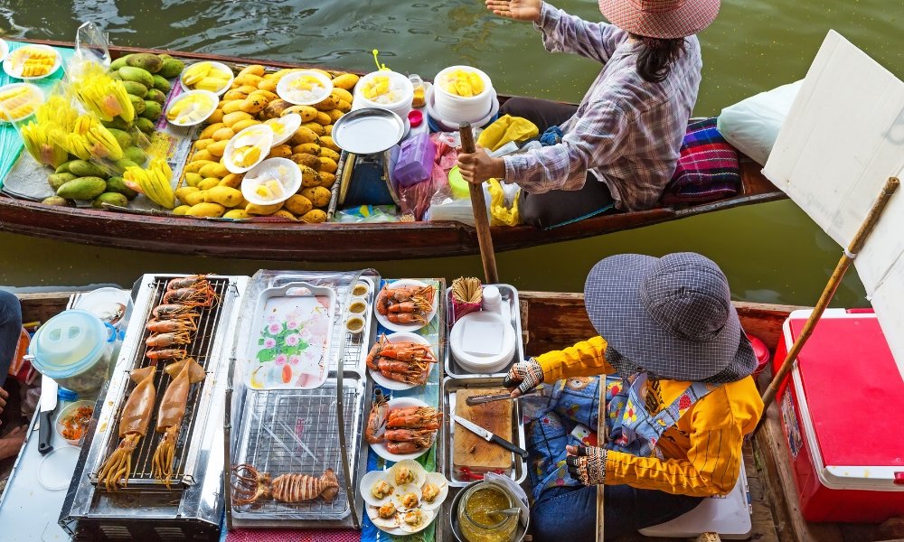 ที่ตลาดน้ำที่คึกคักแห่งนี้ ซึ่งเป็นหนึ่งในสถานที่ท่องเที่ยวที่ดีที่สุดในกรุงเทพฯ มีพ่อค้าแม่ค้าสองคน ที่เที่ยวกรุงเทพ ในเรือให้บริการอาหารทะเลย่าง ผลไม้สด และอาหารอร่อย ๆ — คนหนึ่งเชี่ยวชาญในการขายมะม่วง ในขณะที่อีกคนปรุงอาหารทะเลที่อร่อย.