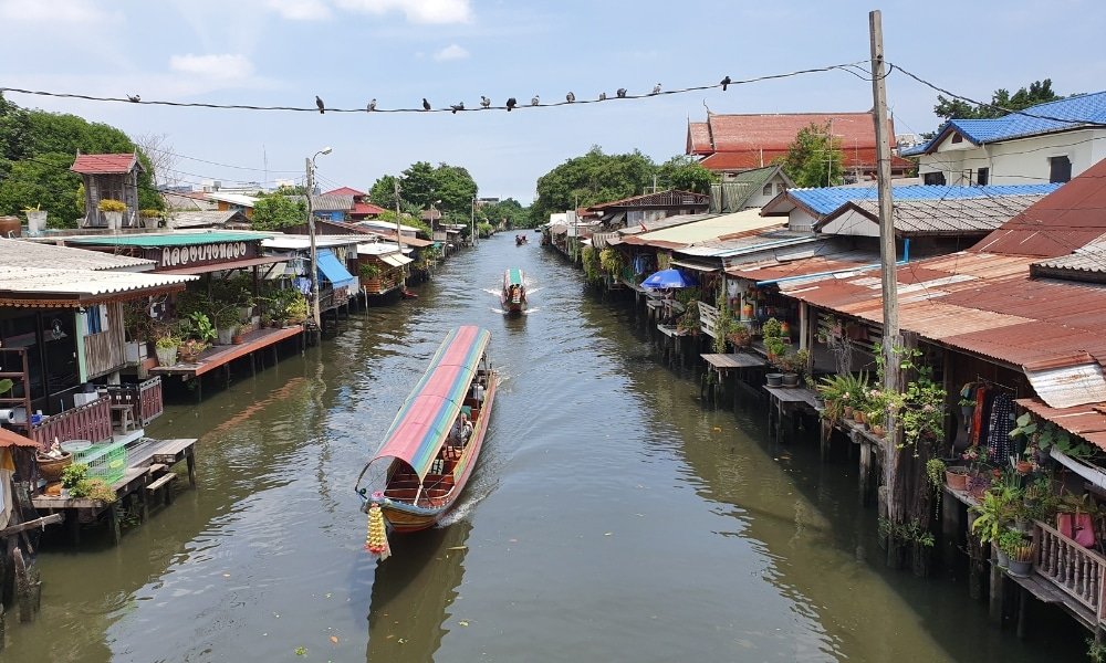 เรือหางยาวแล่นไปตามคลองที่เรียงรายไปด้วยบ้านเรือนที่สร้างบนเสา ที่เที่ยวกรุงเทพ สูง ขณะที่นกหลายตัวเกาะอยู่บนสายไฟเหนือผืนน้ำ—ถ่ายทอดเสน่ห์ของสถานที่ท่องเที่ยวในกรุงเทพฯ.